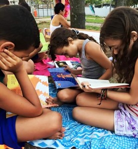 Crianças do projeto leem livros em uma atividade realizada em um parque. Elas estão sentadas na grama, em cima de panos coloridos.